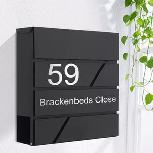 May include: A black, contemporary mailbox displaying the number "59" in silver and the address "Brackenbeds Close." The mailbox has a rectangular form with angled design elements. It is mounted on a white wall.