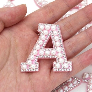 May include: A pink letter "A" decorated with faux pearls and rhinestones. The letter has a border of small, clear rhinestones. The centre of the letter is open, and it is held in a hand.
