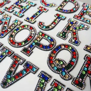 May include: A selection of alphabet letters, each adorned with multi-coloured rhinestones. The letters are outlined in black and feature a variety of jewel-toned colours, including red, green, blue, and yellow. The letters are arranged on a white surface.