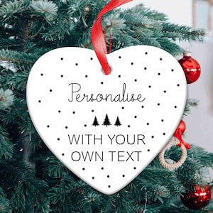 May include: A white heart-shaped Christmas tree decoration with the word "Personalise" in black script. The ornament has a red ribbon for hanging and is decorated with black star-shaped accents and three small Christmas trees. The text "WITH YOUR OWN TEXT" is also included.