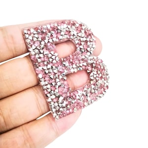 May include: A decorative letter "B" covered in pink and silver rhinestones. The letter is held in a hand, showing the detail of the crystal embellishments. The background is plain white, highlighting the decorative letter. Ideal for crafts.