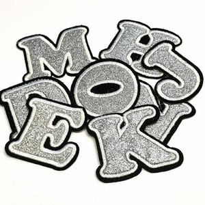 May include: A selection of silver glitter letter patches with black edging. The letters include M, A, J, O, E, K, and D. These iron-on patches are suitable for personalising clothing, bags, or other fabric items.