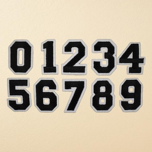May include: A set of black and white embroidered number patches, from zero to nine. Each number features a black central area with a white border, displayed on a cream-coloured backdrop. These patches are suitable for personalising clothing or accessories.