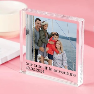 May include: A clear acrylic photo block with a family portrait on a boat. The image shows a man, woman, and two children. Text on the block reads "our cute little adventure" and "28.02.2024". The block is on a pink surface.