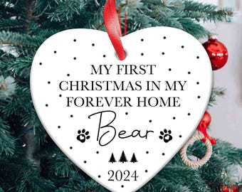 May include: White heart-shaped Christmas ornament with black text that reads "MY FIRST CHRISTMAS IN MY FOREVER HOME Bear 2024". The ornament has a red ribbon for hanging and is decorated with black stars and paw prints.