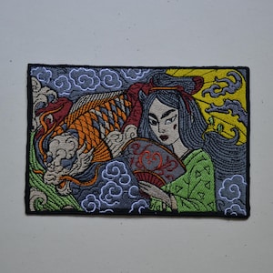 May include: A rectangular embroidered patch featuring a person with long dark hair holding a red and black folding fan. Beside them is a large orange and white koi fish swimming through stylized grey clouds and green waves against a yellow sun.