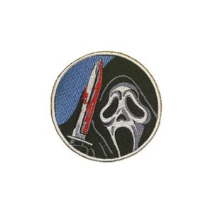 Può includere: Toppa ricamata circolare raffigurante una maschera di Scream e un coltello insanguinato. La toppa ha uno sfondo nero, un cielo blu e un bordo dorato. La maschera è bianca e grigia, e il coltello è argentato con sangue rosso.