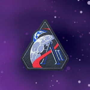 Puede incluir: Parche bordado negro en forma de triángulo, con un diseño de luna con un planeta azul y blanco, una franja roja y la letra "A" en blanco. El parche tiene un borde blanco. El fondo es un degradado púrpura.