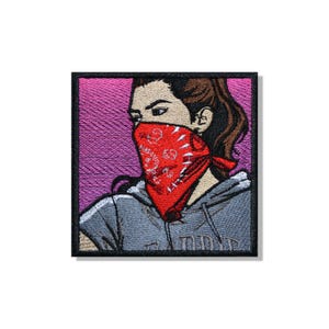 Puede incluir: Un parche bordado cuadrado que muestra a una persona de cabello castaño con una sudadera gris con capucha y un pañuelo rojo brillante con un patrón de cachemira blanco sobre la cara, sobre un fondo degradado de tonos violetas y rosas.