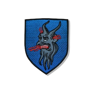 Peut inclure: Un écusson brodé en forme de bouclier avec un fond bleu et une bordure noire. Au centre, une tête de créature grise à cornes avec une bouche grande ouverte montrant des dents blanches acérées et des détails rouges en forme de flammes.