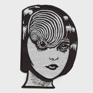 May include: A black and white embroidered patch featuring a stylized face with a large spiral pattern covering the forehead and one eye. The design includes detailed line work for the hair, nose, and lips, with a solid black border edge.
