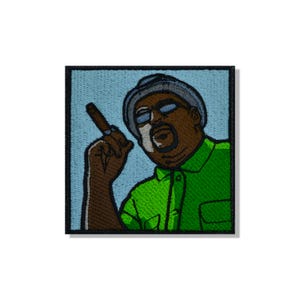 Op de afbeelding: Een vierkante geborduurde patch met een stripfiguur die een blauwe hoed en een felgroen overhemd met kraag draagt. Het personage heeft een donkere huid, draagt een bril en houdt een bruine sigaar vast. De achtergrond is lichtblauw met een zwarte rand.