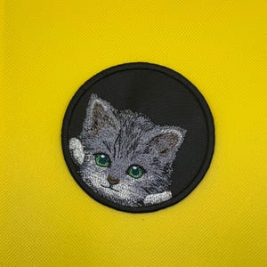 May include: A circular embroidered patch featuring a grey tabby kitten with bright green eyes. The kitten is depicted from the chest up, set against a solid black background within a thick black stitched border, creating a detailed feline portrait.