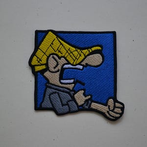 May include: A square embroidered patch featuring a cartoon character with a yellow hat, tan skin, and a grey shirt, set against a solid blue background. The character is shown in profile with an open mouth, pointing forward with a tan hand.