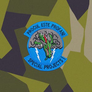 May include: A circular embroidered patch featuring a green dragon head emerging from a grey cloud with white lightning bolts. The blue border contains the text PROCUL ESTE PROFANI at the top and SPECIAL PROJECTS at the bottom in black lettering.