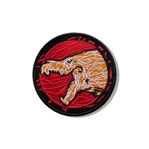 Puede incluir: Un parche bordado circular que presenta una cabeza de criatura estilizada en naranja y blanco con la boca abierta, sobre un fondo rojo sólido. El diseño está encerrado dentro de un borde negro grueso, creando un emblema gráfico de alto contraste.
