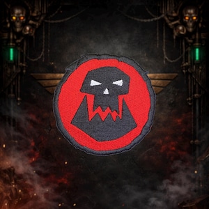 May include: Embroidered patch featuring a black skull with white eyes and teeth, set within a red circle. The patch has a black border and is likely for clothing or accessories. The design is bold and graphic.