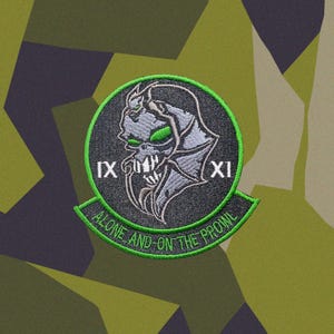 Pode incluir: Um emblema bordado circular apresentando uma criatura cinza semelhante a um crânio com olhos e dentes verde-brilhantes. Os algarismos romanos IX e XI ladeiam a figura central. Uma faixa curva exibe o texto ALONE AND ON THE PROWL.