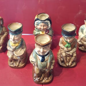 Op de afbeelding: Een verzameling van zeven vintage keramische karakterkruiken. Elke kruik is een gedetailleerd beeldje van een persoon, met een holle bovenkant. De kruiken hebben verschillende kleuren, waaronder bruin, groen en zwart, en zijn gerangschikt op een rode ondergrond.
