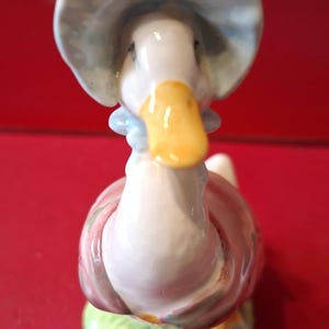 May include: A ceramic figurine of a white duck with a yellow beak, wearing a light blue bonnet and a pink shawl. The duck is standing on a textured green base that resembles grass, with a glossy finish throughout the entire piece.