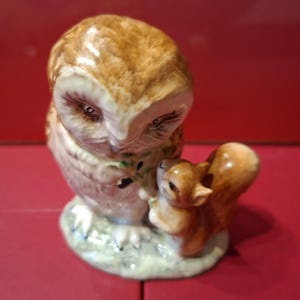 May include: A glazed ceramic figurine featuring a brown and white owl standing next to a small brown squirrel. The owl is looking down at the squirrel, and both animals are positioned on a textured, light-colored base with green accents.