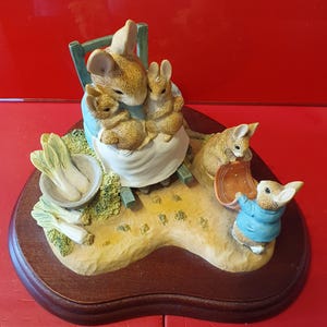 Puede incluir: Figurilla detallada que representa una madre ratón y sus hijos, sentados en una silla verde. La escena incluye un cuenco de verduras y un conejo con una chaqueta azul. La base es una placa de madera marrón oscuro.