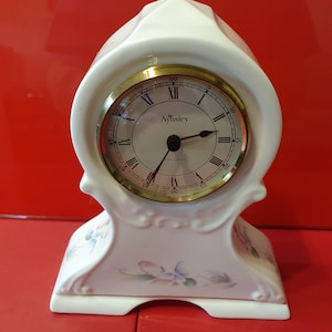 May include: White porcelain clock with gold accents and floral details. The clock face features Roman numerals and the text "Aynsley England".