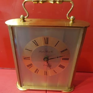 May include: A gold-toned, rectangular travel clock with a handle. The clock face is a light gold color with black Roman numerals and the words "London Clock Co." The clock has a quartz movement.