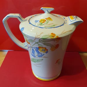 Puede incluir: Una cafetera de cerámica blanca con un diseño floral. Las flores son amarillas, naranjas y azules. La cafetera tiene una tapa con un borde amarillo y azul.