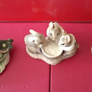 Op de afbeelding: Drie decoratieve konijnenbeeldjes. Eén toont een konijn dat in een peul slaapt, een ander beeldt drie konijnen rond een kom af, en de laatste toont een konijn in een blauw jasje bij een bloempot.
