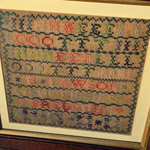 以下が含まれることがあります： ベージュのジュートのような背景に、カラフルなクロスステッチのテキストが施された、額装されたアンティークのニードルポイントサンプラー。テキストには名前と日付が含まれており、金色のフレームに額装されています。ヴィンテージのテキスタイルアート作品です。