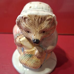 May include: A ceramic figurine of a hedgehog wearing a white bonnet with gold dots, a blue jacket, and yellow trousers. The hedgehog is holding a small yellow rectangular object and a woven basket, standing on a circular white base.