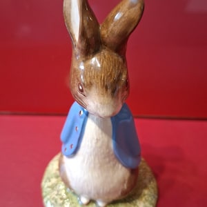 May include: A ceramic figurine of a brown rabbit with long ears, wearing a blue jacket. The rabbit stands on a textured, circular green base. The figure features painted details including whiskers, dark eyes, and a white chest area, with a glossy finish.