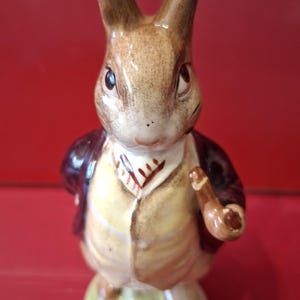 May include: A ceramic figurine of a brown rabbit standing upright on a circular base. The rabbit wears a dark red jacket over a yellow waistcoat and a white cravat, and holds a small brown pipe in one hand with a painted facial expression.