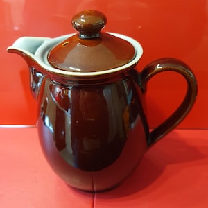 Pode incluir: Uma cafeteira de cerâmica marrom com tampa e alça. A cafeteira tem uma borda branca ao redor da parte superior.