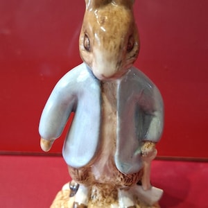 May include: A ceramic figurine of a brown rabbit standing on a textured base. The rabbit has long ears and is wearing a light blue jacket. It is holding a small white shovel in one hand. The figure is painted with detailed fur textures.