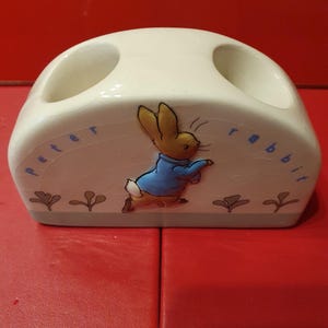 Puede incluir: Portacepillos de cerámica color crema con diseño de Peter Rabbit. El soporte tiene dos aberturas circulares y una ilustración pintada de Peter Rabbit con una chaqueta azul. Las palabras "Peter Rabbit" están escritas en la parte delantera.