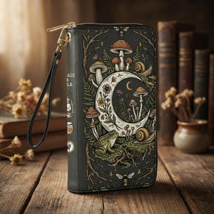 May include: Black wallet with a whimsical forest scene featuring mushrooms, a crescent moon, and a frog. The wallet has a gold zip and wrist strap. The text "AGE & LK" is visible on the side.