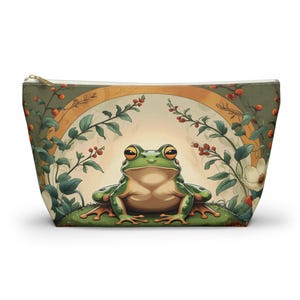 May include: A cosmetic bag with a green frog design. The frog is depicted on a grassy area, surrounded by leaves and a golden arch. The bag has a cream background and a gold zip. The overall design has a whimsical feel.