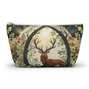 May include: A zippered pouch featuring a detailed illustration of a deer with large antlers, set within a forest scene. The design includes a sunburst, trees, and foliage in shades of brown, green, and cream. The pouch has a gold zipper.