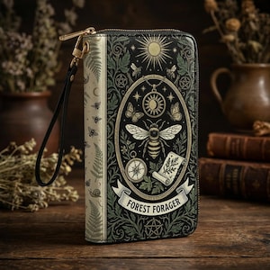 Könnte beinhalten: Eine schwarze und cremefarbene Geldbörse mit goldenem Reißverschluss und Handschlaufe. Die Geldbörse zeigt eine detaillierte Illustration einer Biene, Sonne, Schmetterlingen und die Worte "Forest Forager" auf der Vorderseite. Das Design hat ein Vintage-Botanisches Thema.