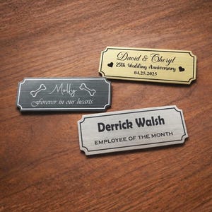 Three engraved metal nameplates displayed on a wooden table: a black pet memorial plaque reading “Molly – Forever in our hearts” with bone icons, a gold “David & Cheryl – 25th Wedding Anniversary – 04.25.2025” plate with heart symbols, and a silver “Derrick Walsh – Employee of the Month” nameplate, all with decorative borders.