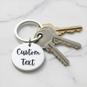 May include: A silver keychain with a round pendant that reads "Custom Text" in a black, cursive font. The keychain is attached to a silver ring with three keys. The background is a white marble surface.
