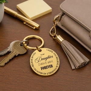 Gold round engraved keychain attached to a set of keys on a wooden desk, reading “To my Daughter I love you forever,” with a circular message around the edge. Nearby are a beige purse with a tassel, a gold pen, sticky notes, and a small potted plant.