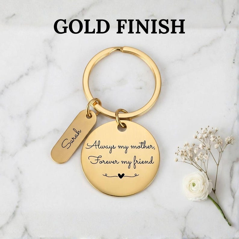 Gold finish personalized keychain for mom with engraved message “Always my mother, forever my friend” and custom name tag, displayed on a marble background with delicate white flowers.