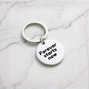 K&ouml;nnte beinhalten: Ein silberner Schl&uuml;sselanh&auml;nger mit einem runden Anh&auml;nger. Auf dem Anh&auml;nger steht in schwarzer Schrift "Forever starts now". Der Schl&uuml;sselanh&auml;nger und der Anh&auml;nger sind aus gl&auml;nzendem Metall gefertigt.