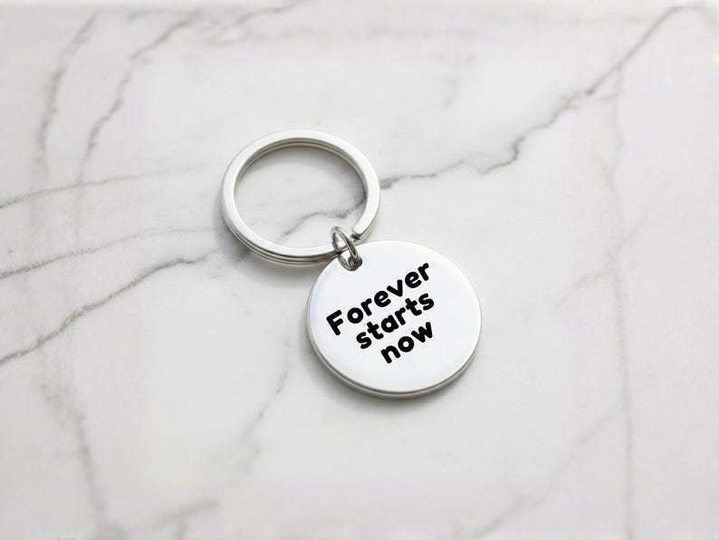 K&ouml;nnte beinhalten: Ein silberner Schl&uuml;sselanh&auml;nger mit einem runden Anh&auml;nger. Auf dem Anh&auml;nger steht in schwarzer Schrift "Forever starts now". Der Schl&uuml;sselanh&auml;nger und der Anh&auml;nger sind aus gl&auml;nzendem Metall gefertigt.