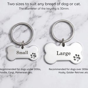 Custom silver pet ID tags for dogs showing small and large size options. Personalized bone-shaped engraved dog name tags with paw print detail on marble.