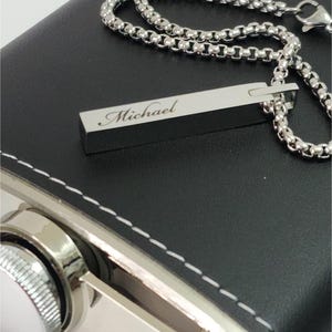 A close-up photograph of a rectangular silver bar pendant necklace resting on a leather-wrapped metal hip flask.