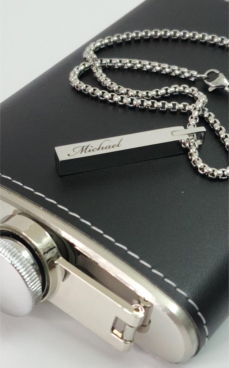 A close-up photograph of a rectangular silver bar pendant necklace resting on a leather-wrapped metal hip flask.